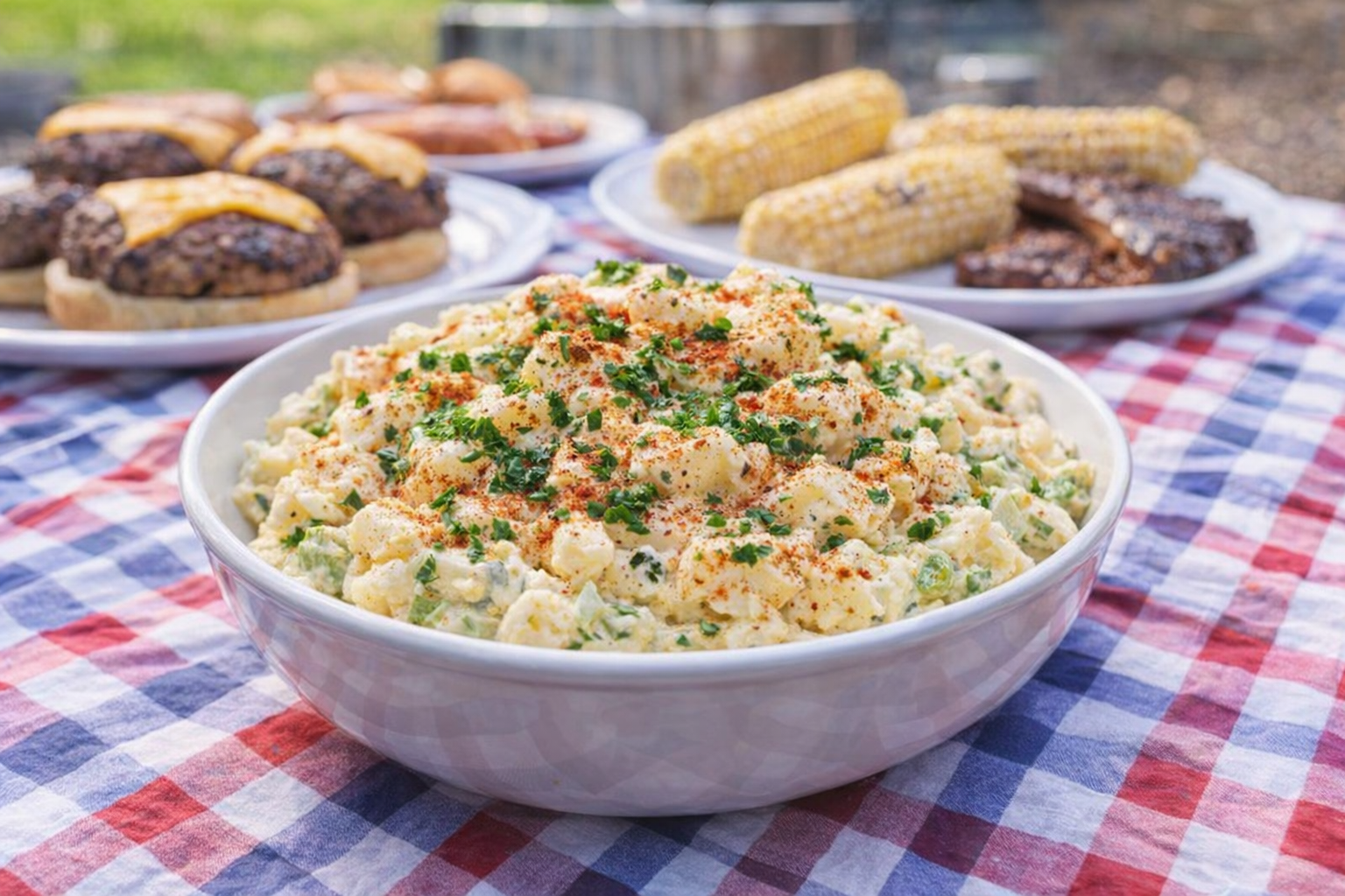 A high-angle shot of a bowl filled with creamy American potato salad, garnished with paprika and fresh parsley, on a picnic blanket with barbecue fare in the background, bright daylight